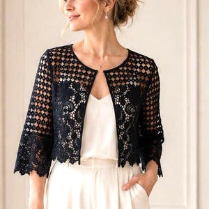 May include: A black lace bolero jacket with a geometric pattern and three-quarter sleeves, worn over a white camisole and high-waisted white wide-leg trousers. A small, rectangular beige clutch is held in one hand, and pointed-toe heels are worn.