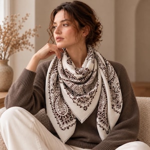Op de afbeelding: Een vierkante sjaal met een crèmekleurige basis en een donkerbruin, ingewikkeld paisley- en bloemenpatroon. De sjaal heeft een gefranjerde rand en is om de nek gedrapeerd, met een symmetrisch ontwerp met gedetailleerde randen en motieven.