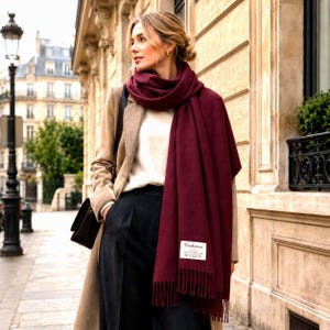 Peut inclure: Une longue écharpe bordeaux profond avec un ourlet à franges et une étiquette rectangulaire blanche portant l'inscription Cashmere. L'écharpe est drapée autour du cou et sur les épaules, portée avec un manteau beige, un haut blanc et un pantalon sombre.