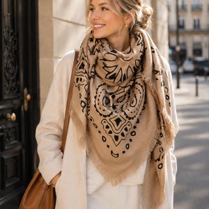 May include: A large, square, beige scarf featuring a bold black paisley and geometric print with frayed edges. The scarf is draped around the neck and shoulders, showcasing a soft, textured fabric construction with a distinct pattern.