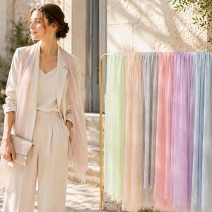 Può includere: Una collezione di sciarpe rettangolari leggere in tonalità pastello verde menta, pesca, grigio, rosa, lavanda e azzurro, esposte su un appendiabiti in metallo dorato, accanto a una persona che indossa un blazer color crema, pantaloni e sciarpa.