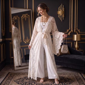 May include: A floor-length cream lace duster with long bell sleeves and a tie waist, worn over a white sleeveless top and wide-leg trousers. The outfit is paired with a small, scalloped cream handbag and a multi-layered gold pendant necklace.