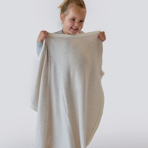 Notre jolie Anaëlle tient dans ses mains la magnifique couverture Botiboty 100% cachemire beige pour nous montrer comme elle est grande. elle mesure 80x100cm avec des fils luxueux soigneusement choisis par nos soins. fabrication made in Madagascar