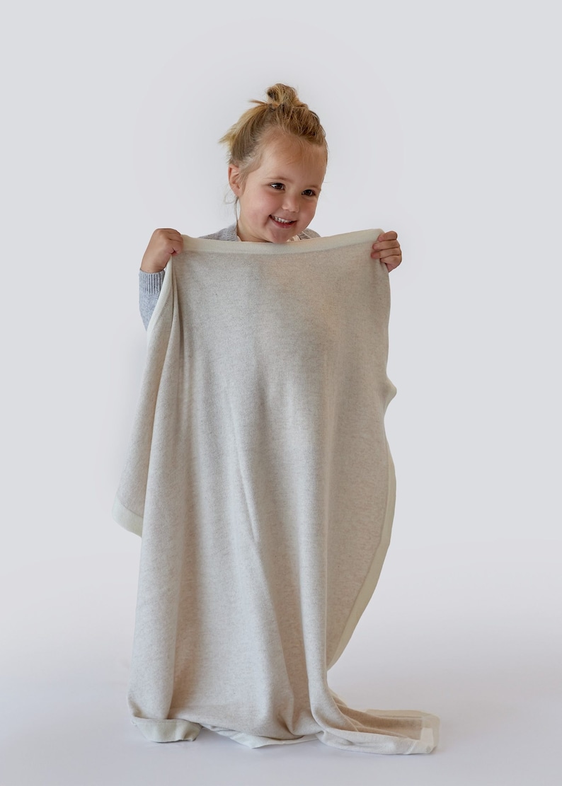 Notre jolie Anaëlle tient dans ses mains la magnifique couverture Botiboty 100% cachemire beige pour nous montrer comme elle est grande. elle mesure 80x100cm avec des fils luxueux soigneusement choisis par nos soins. fabrication made in Madagascar
