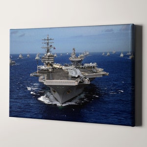 May include: A large gray aircraft carrier sails on a blue ocean with a fleet of smaller ships behind it. The carrier has a long flight deck and a large island structure.