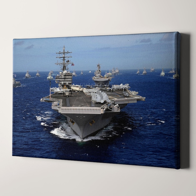May include: A large gray aircraft carrier sails on a blue ocean with a fleet of smaller ships behind it. The carrier has a long flight deck and a large island structure.