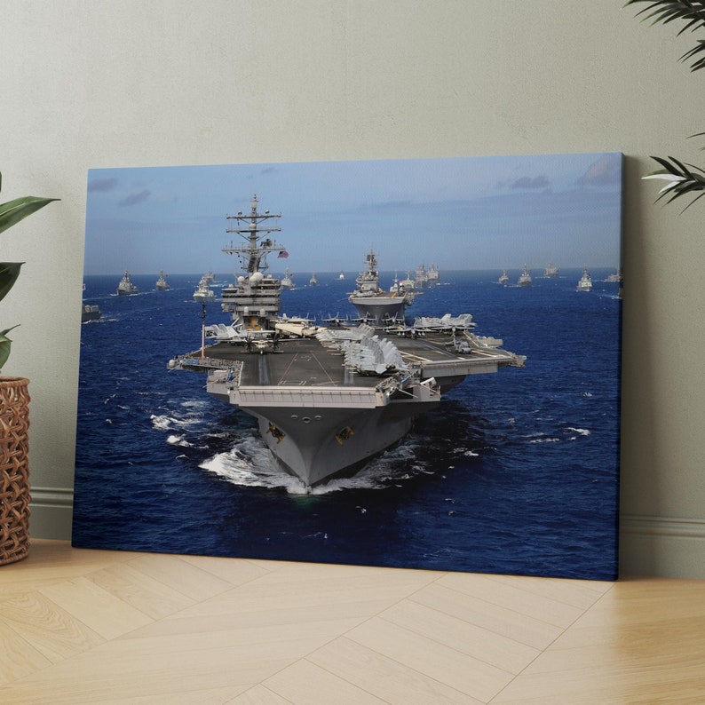May include: A large gray aircraft carrier with a flight deck and several smaller ships surrounding it. The carrier is in the middle of the ocean, with blue water and a clear sky in the background.