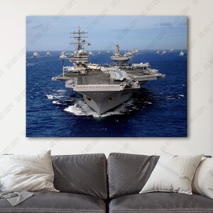 May include: An aerial view of a large aircraft carrier sailing on a blue ocean. The carrier is gray and has a long flight deck. Several smaller ships are visible in the background.