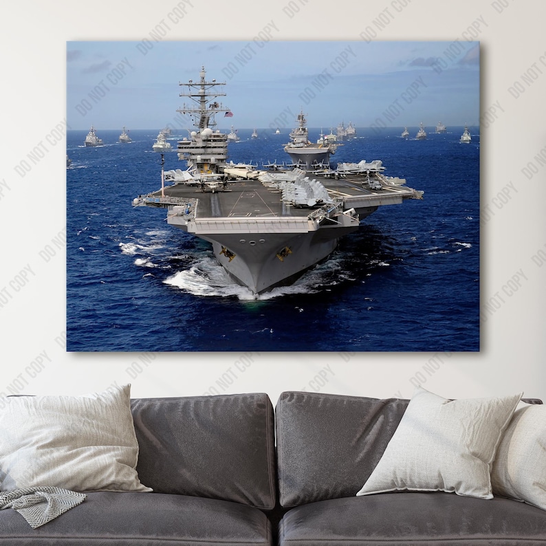 May include: An aerial view of a large aircraft carrier sailing on a blue ocean. The carrier is gray and has a long flight deck. Several smaller ships are visible in the background.