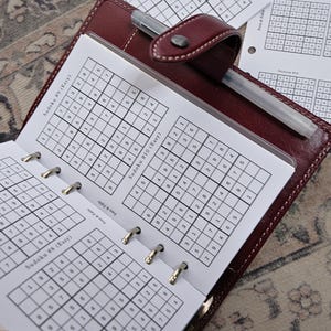 May include: A brown leather planner open to Sudoku puzzles. The pages display printed Sudoku grids with numbers. A silver pen rests inside the planner. Additional Sudoku sheets are visible in the background. The planner is on a patterned surface.