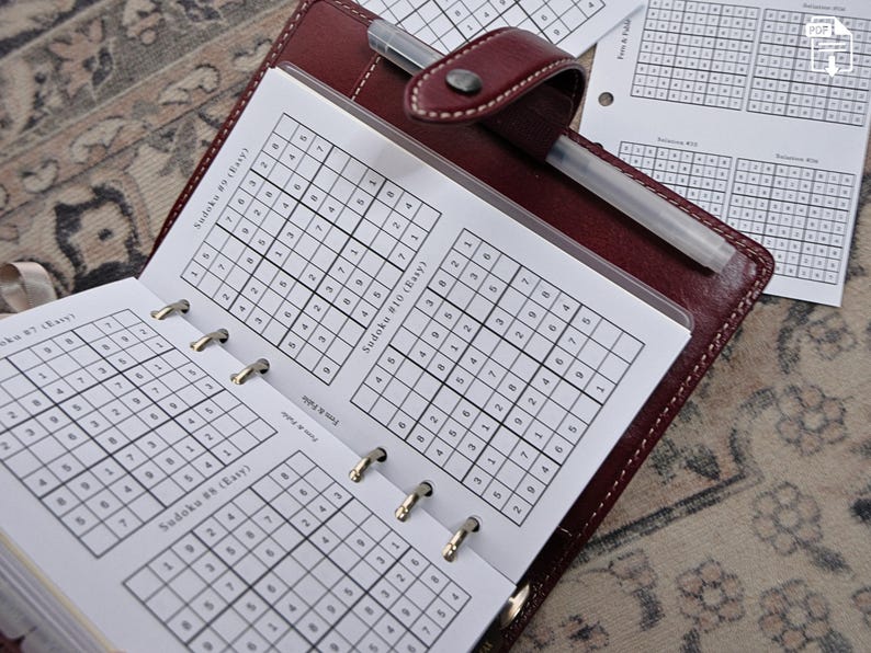 May include: A brown leather planner open to Sudoku puzzles. The pages display printed Sudoku grids with numbers. A silver pen rests inside the planner. Additional Sudoku sheets are visible in the background. The planner is on a patterned surface.
