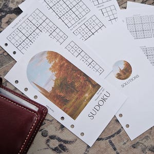 May include: A collection of printed Sudoku puzzle sheets with varying difficulty levels, including "Easy," "Medium," and "Hard." The sheets feature Sudoku grids and are accompanied by a burgundy leather planner. One sheet has a landscape painting and the word "SUDOKU."