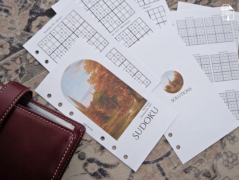 May include: A collection of printed Sudoku puzzle sheets with varying difficulty levels, including "Easy," "Medium," and "Hard." The sheets feature Sudoku grids and are accompanied by a burgundy leather planner. One sheet has a landscape painting and the word "SUDOKU."