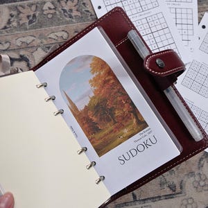 May include: Burgundy leather planner with a pen, open to a page with a landscape print and the word "SUDOKU". Additional sudoku puzzles are visible on loose-leaf paper.