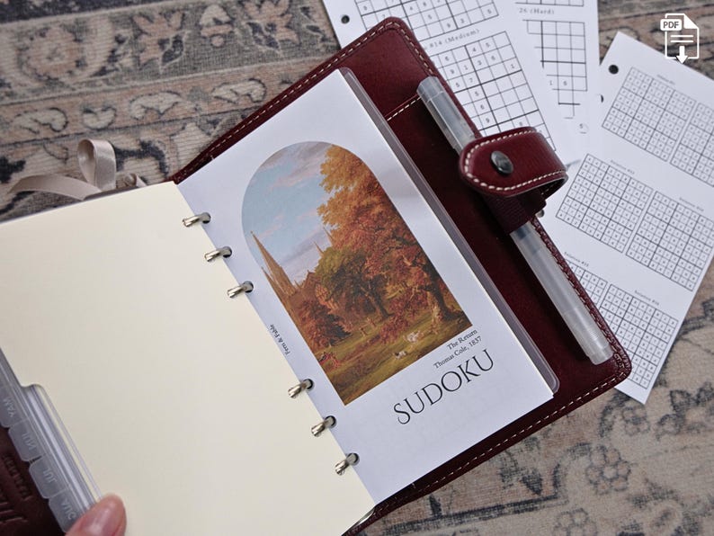 May include: Burgundy leather planner with a pen, open to a page with a landscape print and the word "SUDOKU". Additional sudoku puzzles are visible on loose-leaf paper.