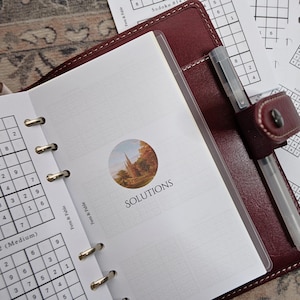 May include: Open burgundy leather planner with a pen and Sudoku puzzles. The planner is open to a page with the word "SOLUTIONS" and a small landscape image. Additional Sudoku puzzles are visible.