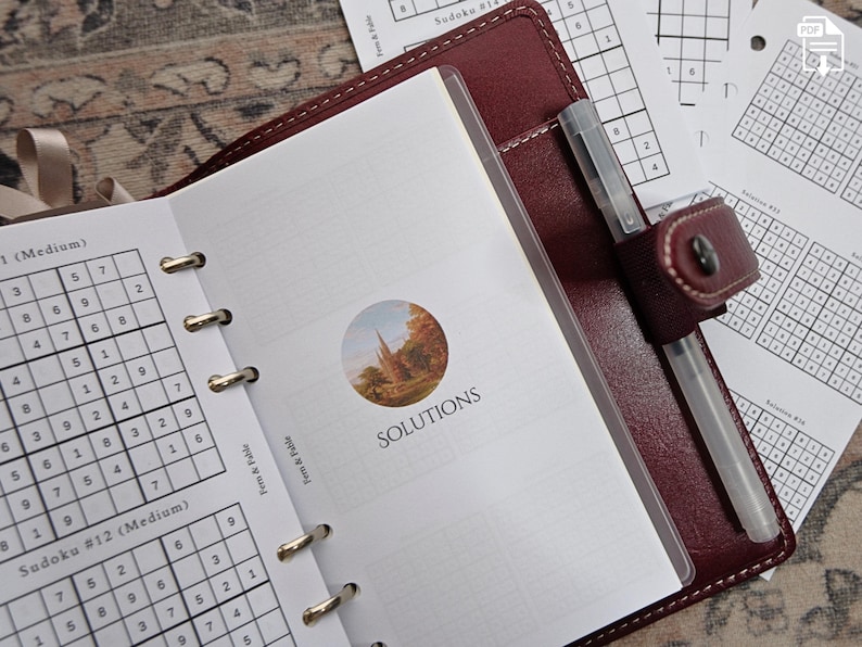 May include: Open burgundy leather planner with a pen and Sudoku puzzles. The planner is open to a page with the word "SOLUTIONS" and a small landscape image. Additional Sudoku puzzles are visible.