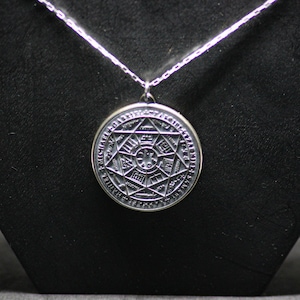 Puede incluir: Un collar de plata con un colgante redondo que presenta un dise&ntilde;o detallado e intrincado de una estrella dentro de un c&iacute;rculo. El colgante est&aacute; rodeado por una inscripci&oacute;n circular.