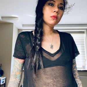 May include: A woman with long black hair braided to the side, wearing a black sheer top with a silver necklace. She has tattoos on both arms.