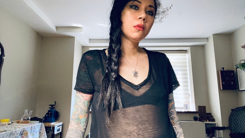 May include: A woman with long black hair braided to the side, wearing a black sheer top with a silver necklace. She has tattoos on both arms.