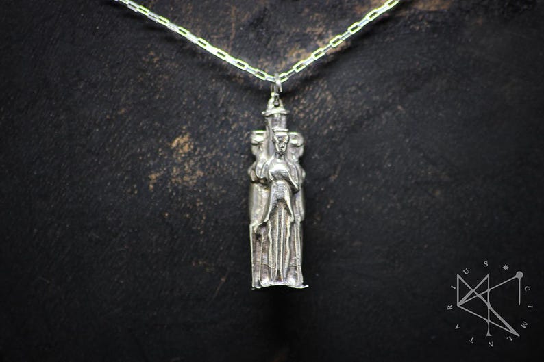 May include: Silver pendant necklace featuring a detailed sculpture of three figures standing close together. The figures are stylized and appear to be wearing robes.