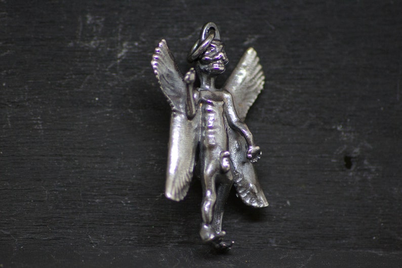 May include: A silver pendant featuring a skeletal angel with outstretched wings. The angel is depicted with a halo and a skeletal face.
