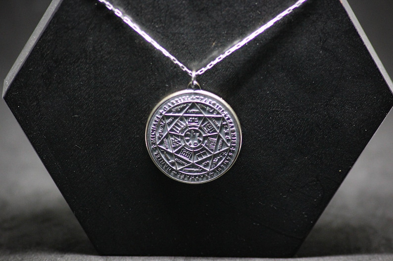 Puede incluir: Un collar de plata con un colgante redondo que presenta un dise&ntilde;o detallado e intrincado de una estrella dentro de un c&iacute;rculo. El colgante est&aacute; rodeado por una inscripci&oacute;n circular.