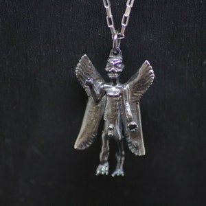 May include: A silver pendant necklace featuring a winged demon figure with a skeletal face. The pendant is detailed and has a textured finish.