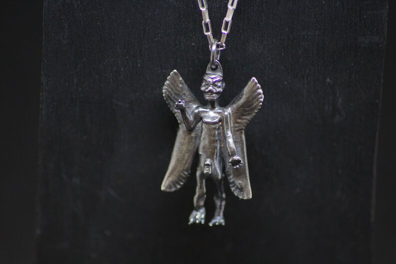May include: A silver pendant necklace featuring a winged demon figure with a skeletal face. The pendant is detailed and has a textured finish.