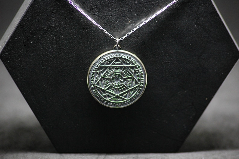 Puede incluir: Un collar de plata con un colgante redondo que presenta un dise&ntilde;o detallado e intrincado de una estrella con un c&iacute;rculo en el centro y texto alrededor del per&iacute;metro.