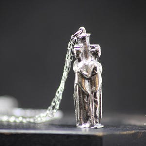 May include: A silver pendant necklace featuring a detailed sculpture of three figures standing close together. The figures are stylized and appear to be embracing. The necklace is on a silver chain.
