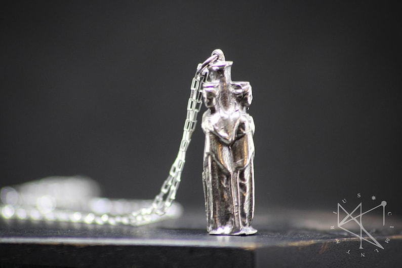 May include: A silver pendant necklace featuring a detailed sculpture of three figures standing close together. The figures are stylized and appear to be embracing. The necklace is on a silver chain.