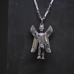 May include: Silver pendant necklace featuring a detailed winged figure with a human-like form. The figure has a long, pointed head and outstretched arms.