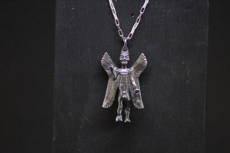 May include: Silver pendant necklace featuring a detailed winged figure with a human-like form. The figure has a long, pointed head and outstretched arms.