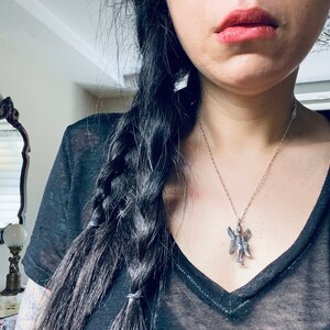 May include: A woman with long black hair braided down her back is wearing a black v-neck t-shirt and a silver necklace with a fairy pendant.