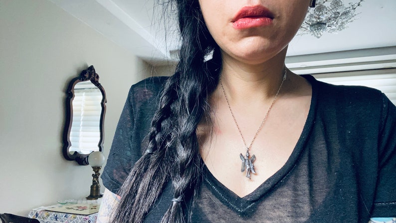 May include: A woman with long black hair braided down her back is wearing a black v-neck t-shirt and a silver necklace with a fairy pendant.