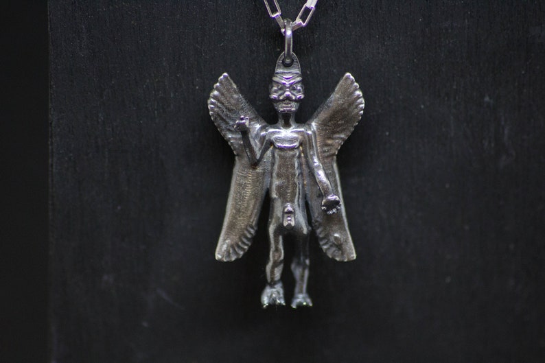 May include: Silver pendant necklace featuring a detailed winged figure with a human-like form. The figure has a pointed head and appears to be wearing a crown.