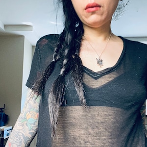 May include: A woman wearing a black sheer top with a v-neck and a silver necklace. Her hair is in a braid and she has a tattoo on her arm.