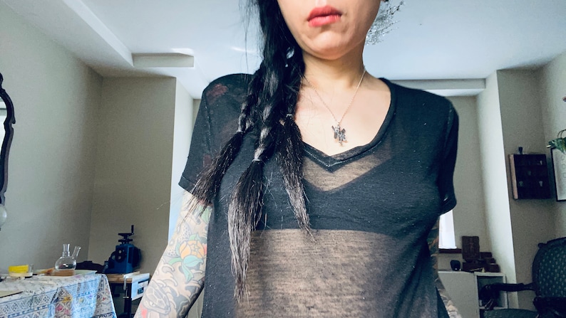 May include: A woman wearing a black sheer top with a v-neck and a silver necklace. Her hair is in a braid and she has a tattoo on her arm.