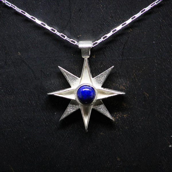 Puede incluir: Un colgante de estrella de ocho puntas de plata con una piedra preciosa de lapislázuli azul en el centro, colgando de una cadena de plata.