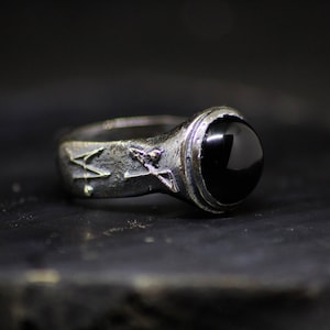 Peut inclure: Une bague en argent avec une pierre précieuse noire sertie dans un sertissage à bélière. La bande de la bague est texturée et présente un motif de flèches.