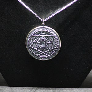 Puede incluir: Un collar con un colgante de plata en forma de c&iacute;rculo adornado con un dise&ntilde;o geom&eacute;trico complejo y una inscripci&oacute;n. El colgante est&aacute; rodeado por un c&iacute;rculo de texto.