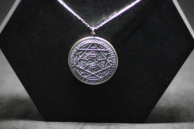 Puede incluir: Un collar con un colgante de plata en forma de c&iacute;rculo adornado con un dise&ntilde;o geom&eacute;trico complejo y una inscripci&oacute;n. El colgante est&aacute; rodeado por un c&iacute;rculo de texto.