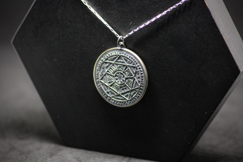 Puede incluir: Un collar de plata con un colgante redondo que presenta un dise&ntilde;o detallado e intrincado de una estrella dentro de un c&iacute;rculo. El colgante est&aacute; grabado con s&iacute;mbolos y caracteres.
