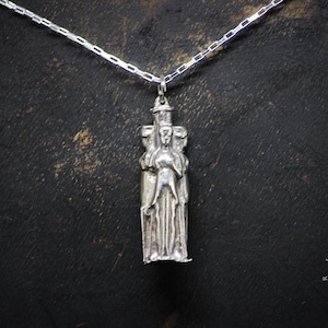 May include: Silver pendant necklace featuring a detailed sculpture of three figures standing together. The figures are stylized and appear to be wearing robes. The pendant is attached to a silver chain.