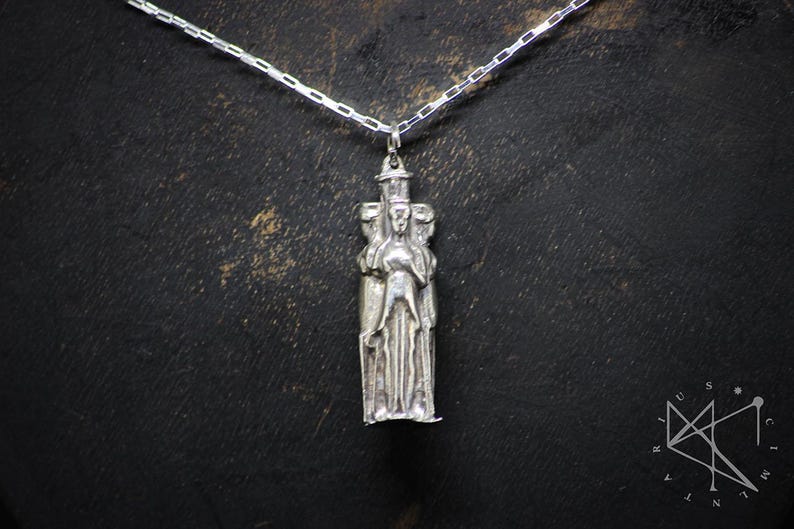 May include: Silver pendant necklace featuring a detailed sculpture of three figures standing together. The figures are stylized and appear to be wearing robes. The pendant is attached to a silver chain.