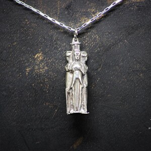May include: Silver pendant necklace featuring a detailed sculpture of three figures standing together. The figures are wearing long robes and have intricate facial features.
