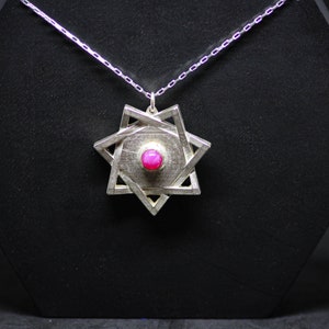 Puede incluir: Un collar con colgante de plata con un diseño de Estrella de David y una gema rosa central. El colgante cuelga de una delicada cadena de plata. El collar se exhibe sobre un fondo negro.