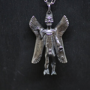 May include: A silver pendant featuring a winged, humanoid figure with a skeletal face. The figure has outstretched arms and appears to be in a standing position.