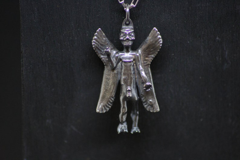 May include: A silver pendant featuring a winged, humanoid figure with a skeletal face. The figure has outstretched arms and appears to be in a standing position.